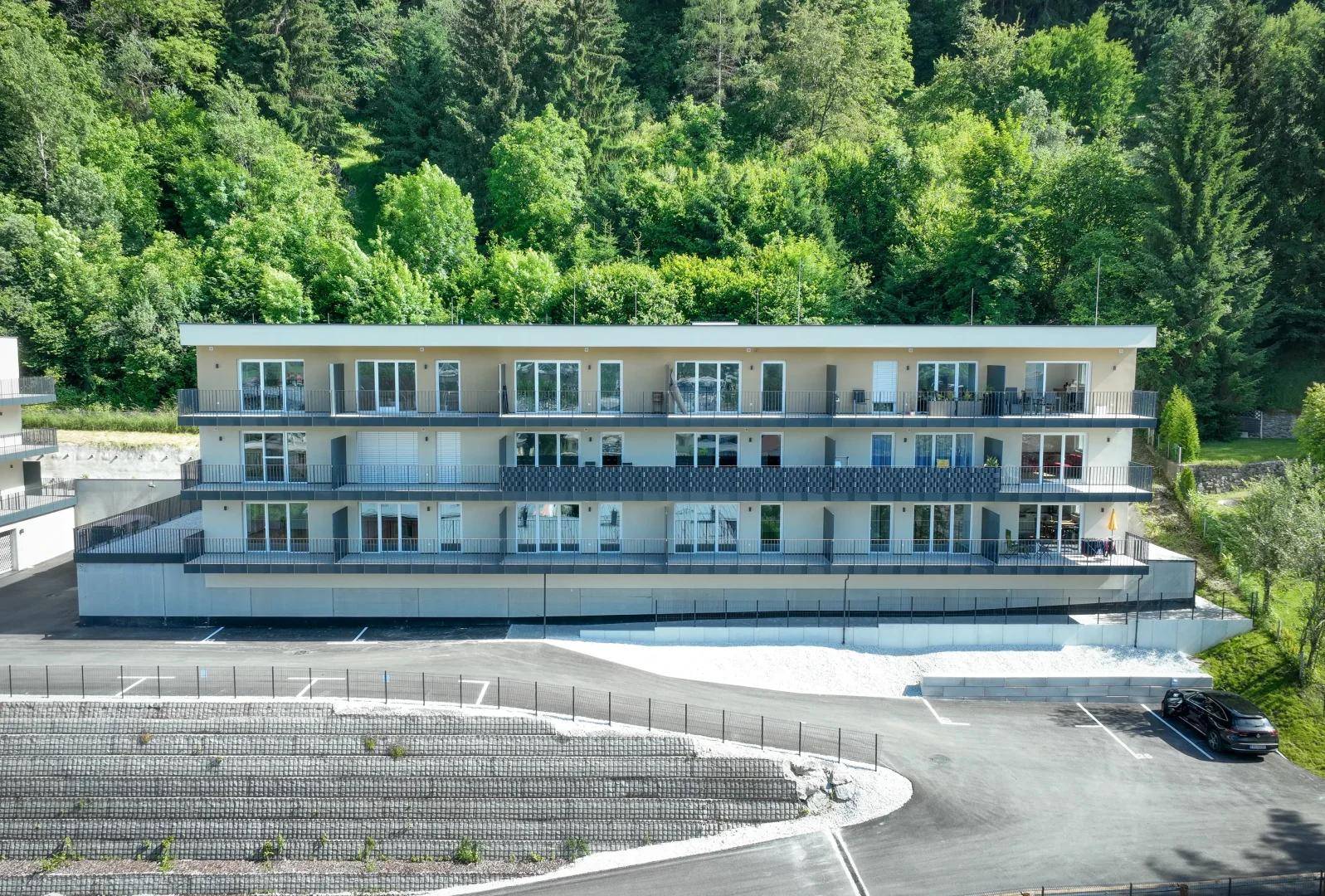 Nahaufnahme eines modernen Apartmentgebäudes mit mehreren Balkonen und großen Fenstern vor bewaldetem Hang.