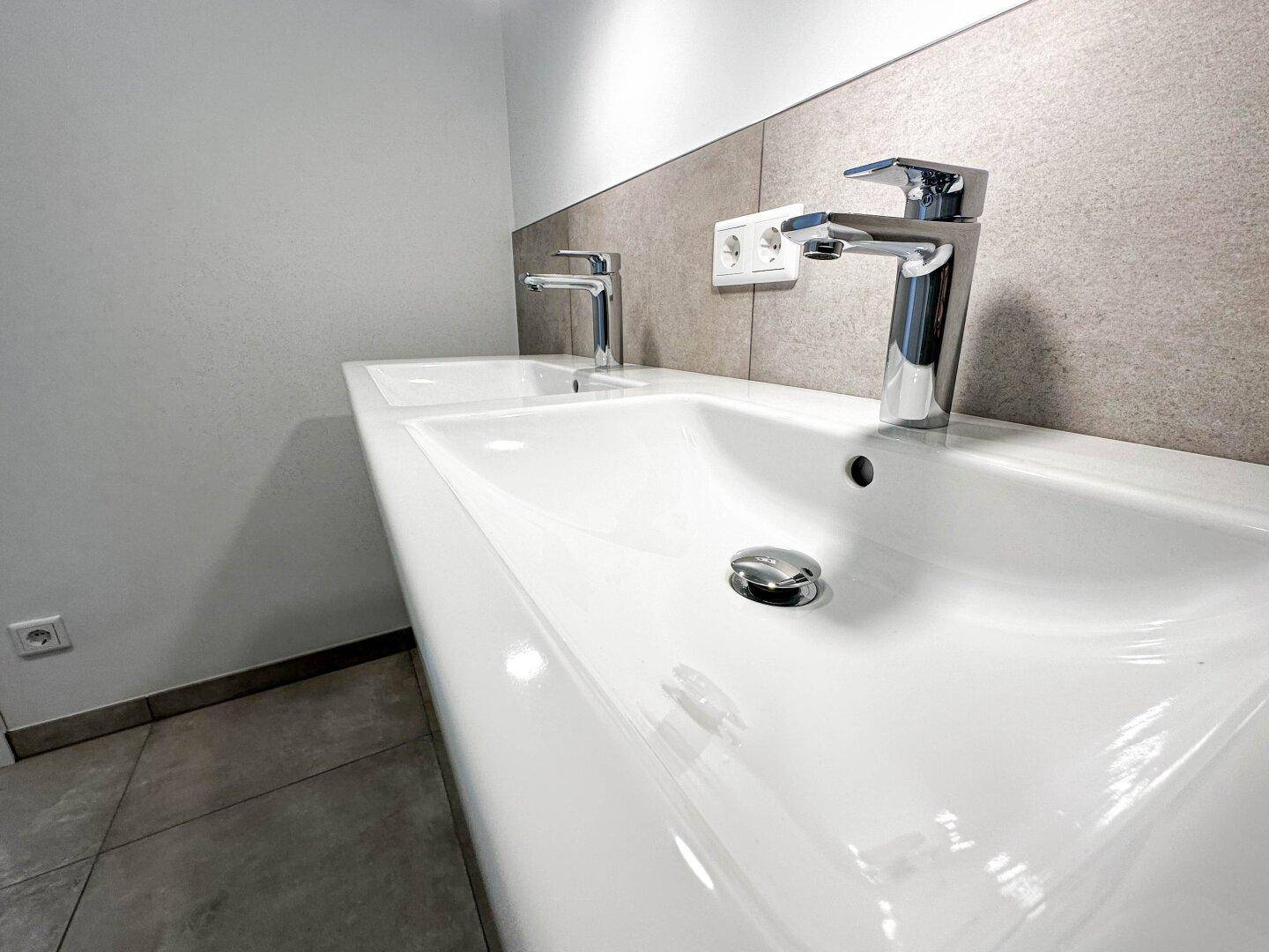 Modernes Badezimmer mit Doppelwaschbecken und eleganten Armaturen auf grauen Fliesen.