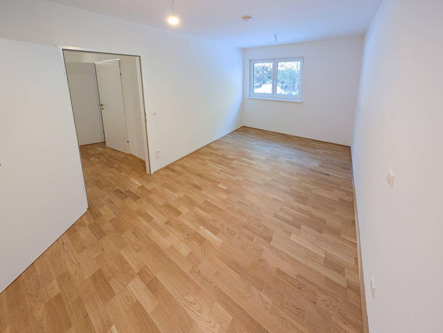 Helles Schlafzimmer mit Parkettboden, weißen Wänden und einem Fenster mit Blick ins Grüne.