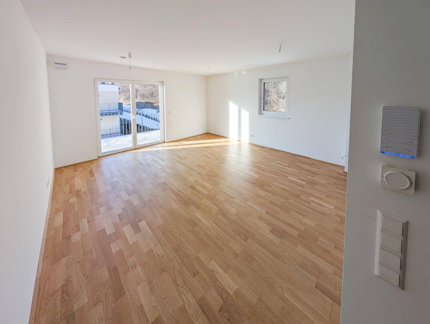 Heller, leerer Wohnraum mit Parkettboden, weißen Wänden und Zugang zum Balkon.