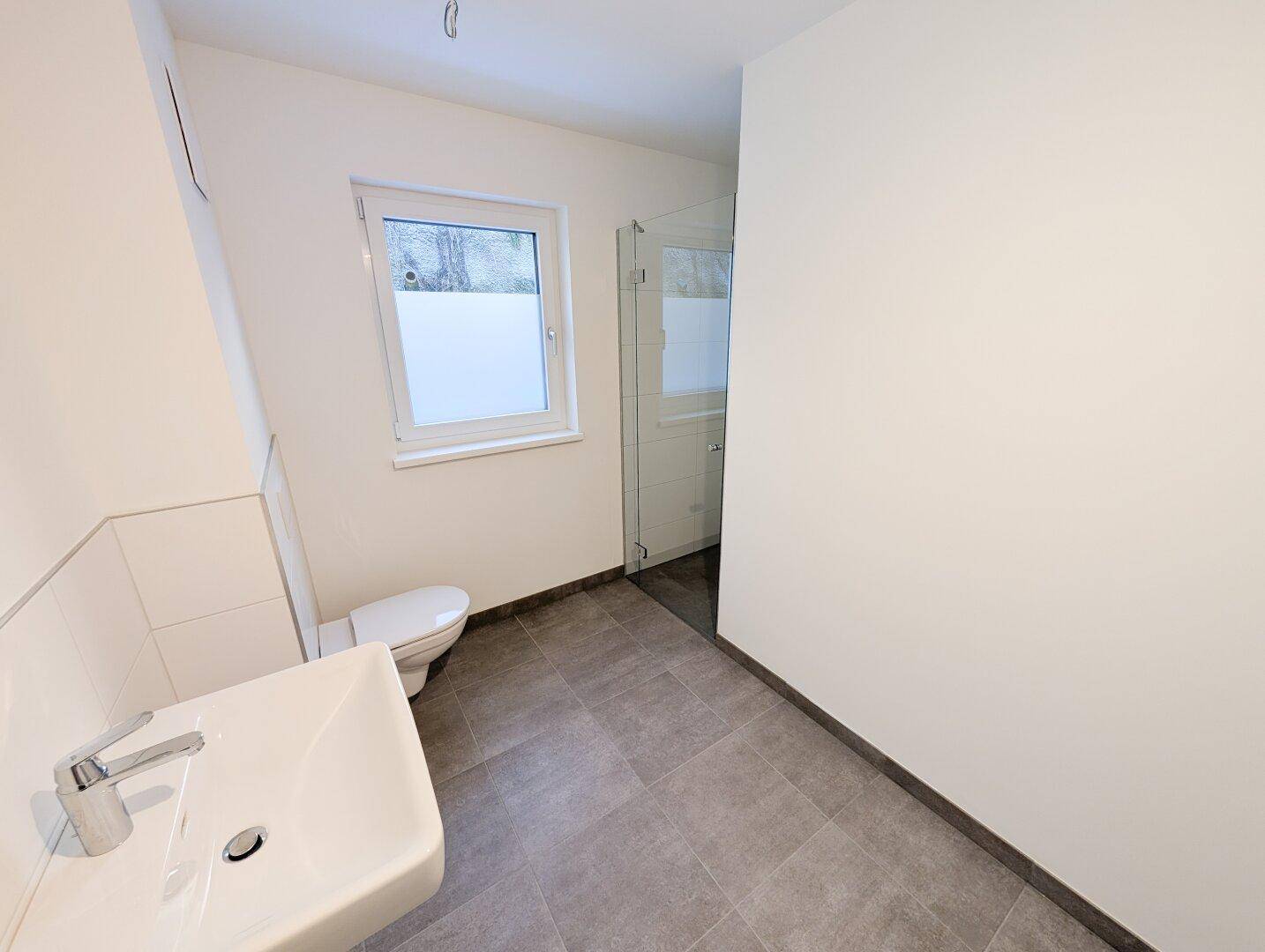 Modernes Badezimmer mit ebenerdiger Dusche, WC, Waschbecken und Fenster für Tageslicht.