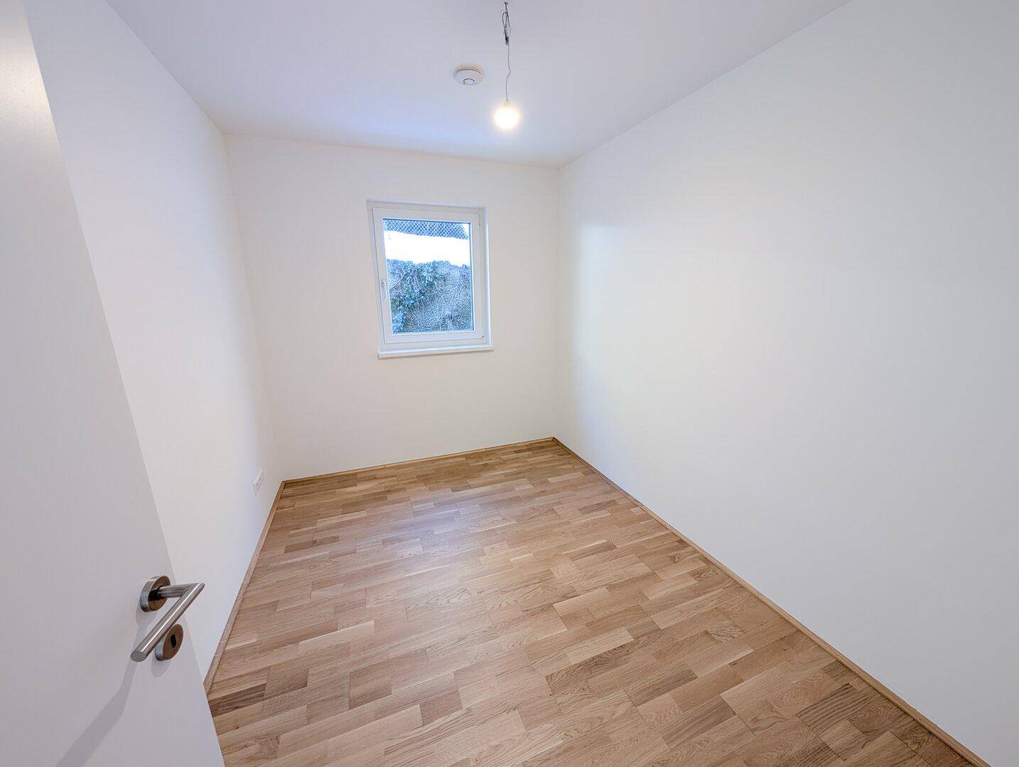 Helles, leeres Zimmer mit Holzboden und einem Fenster, geeignet als Schlaf- oder Kinderzimmer.