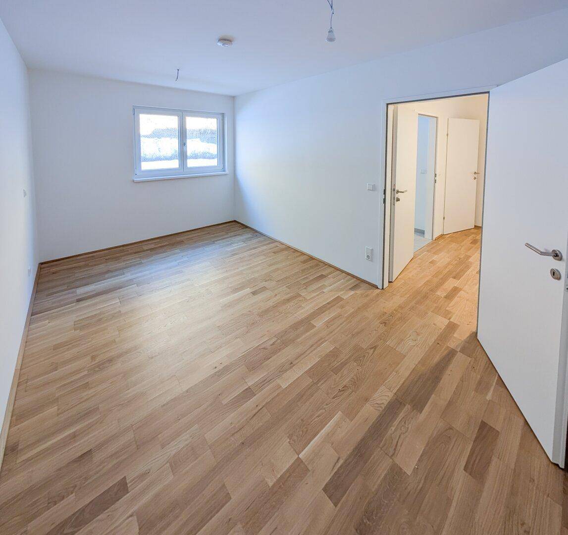 Geräumiges, helles Zimmer mit Holzboden und Fenster, bietet Zugang zu einem Flur.