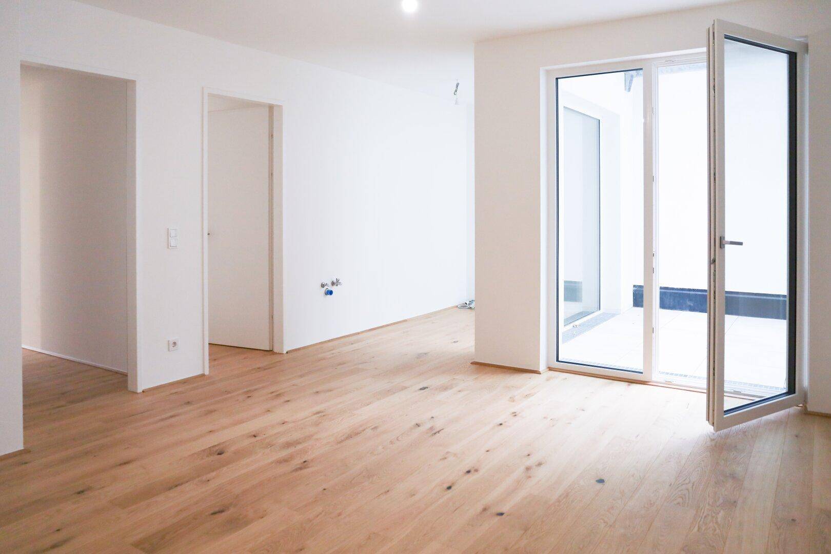 Heller, leerer Raum mit neuem Holzboden und großen Fenstertüren, die viel Licht hereinlassen.