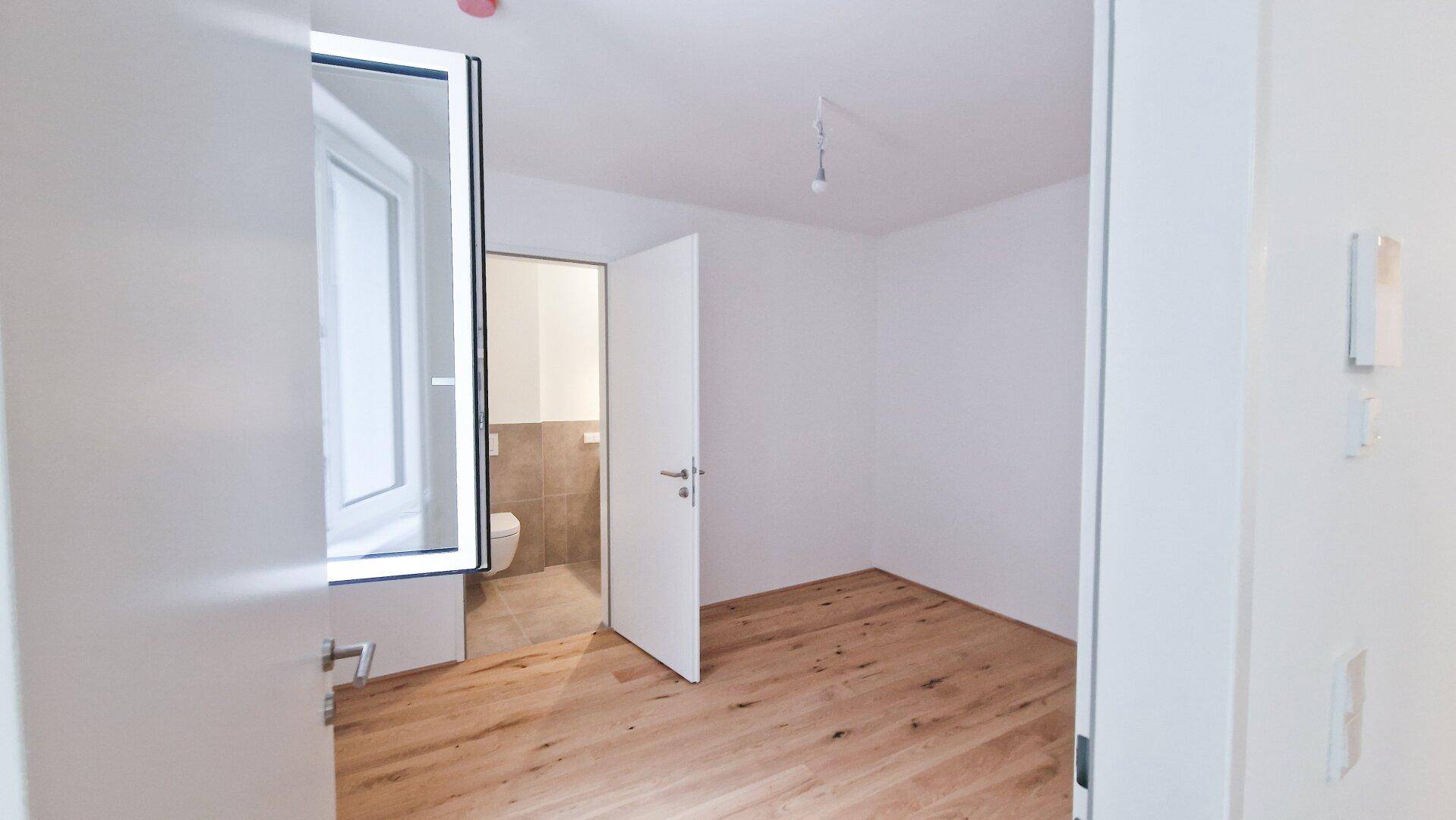 Leerer Raum mit Holzboden und Blick durch eine offene Tür in ein angrenzendes Badezimmer.