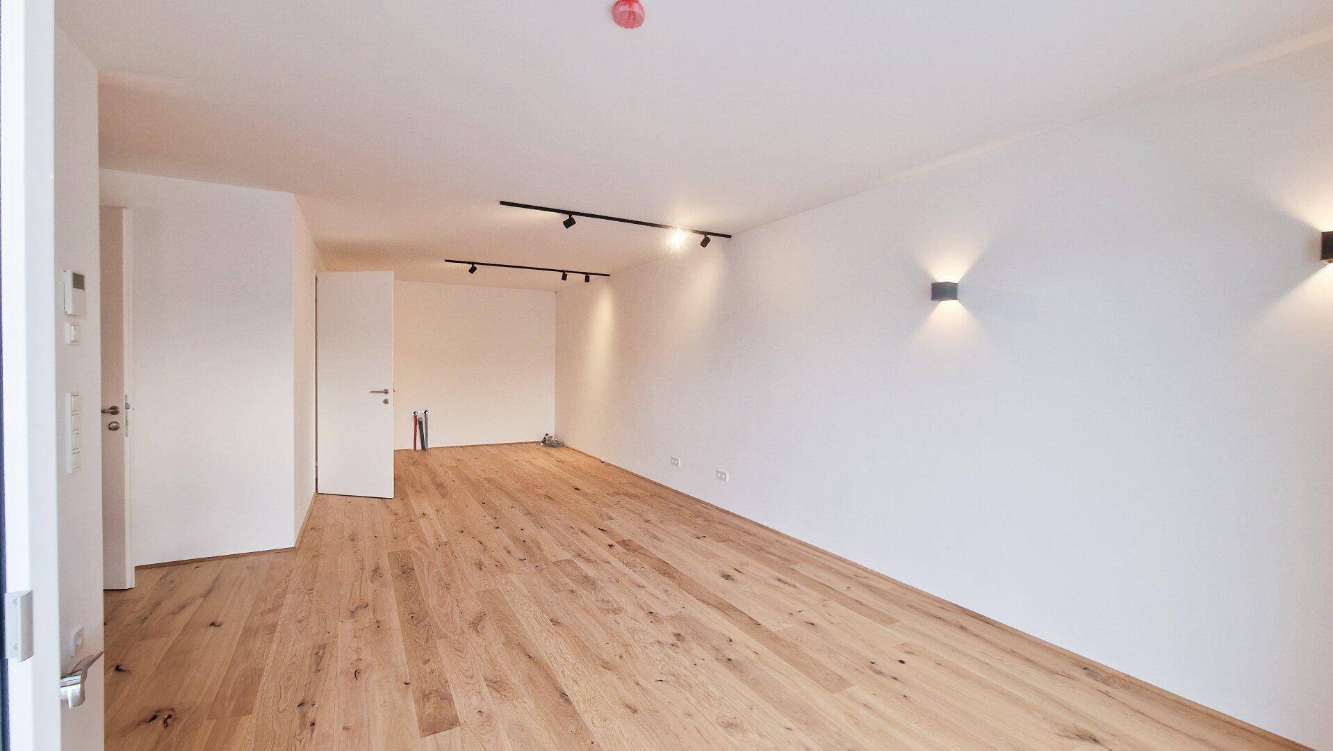 Geräumiger, leerer Wohnbereich mit Holzboden, modernen Wandleuchten und viel natürlichem Licht.