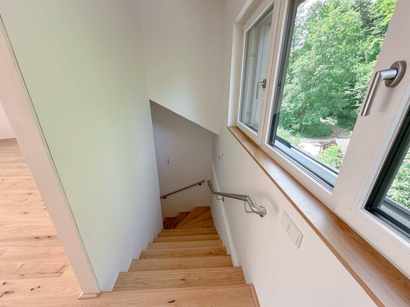 Holztreppe mit Handlauf und Fenster, das einen Blick ins Grüne bietet.