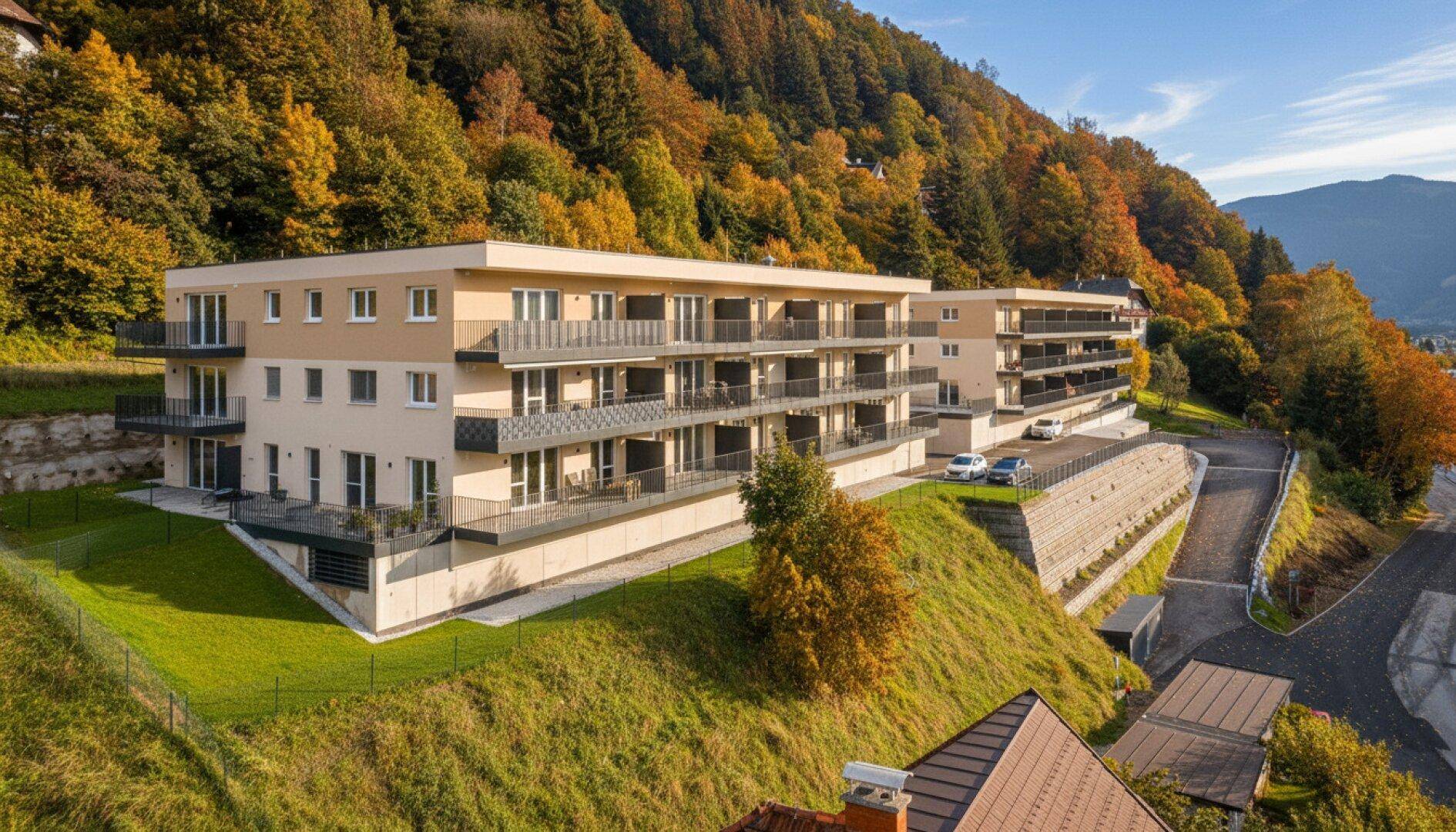 Modernes Apartmentgebäude am Hang, umgeben von herbstlichen Bäumen und Natur.