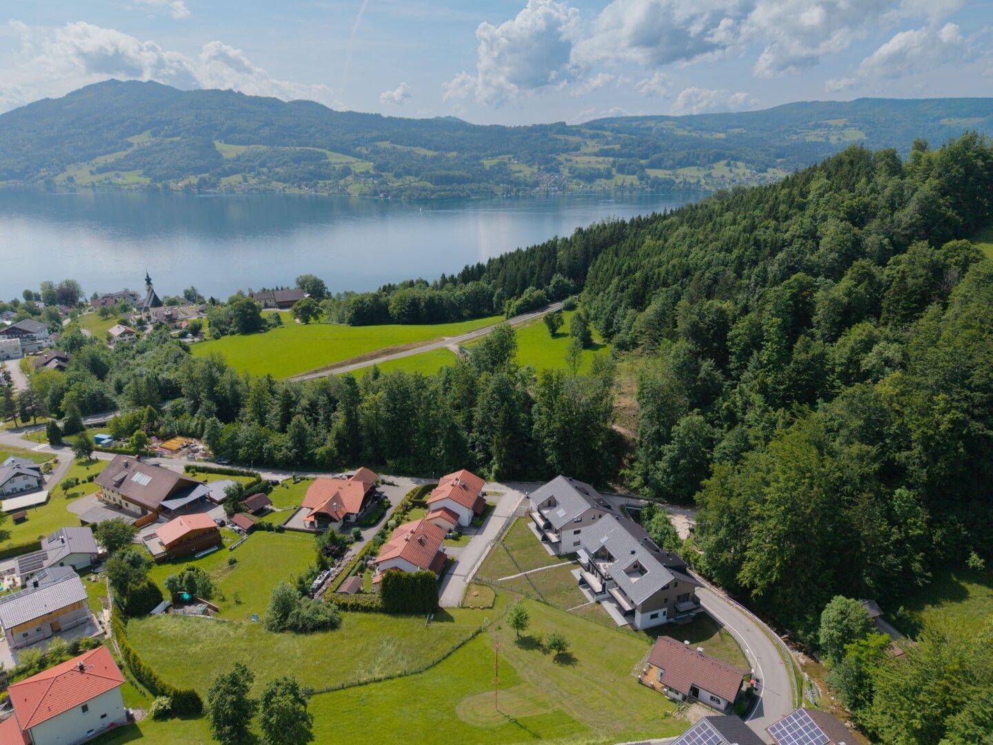 Panoramablick aus der Vogelperspektive auf eine Wohnsiedlung am Seeufer mit umliegenden Bergen und Wäldern.
