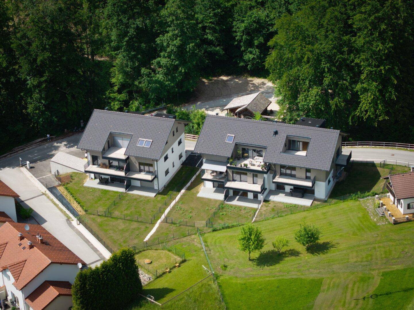 Zwei moderne Neubauten mit Satteldächern, Balkonen und großen Fenstern, umgeben von viel Grün.