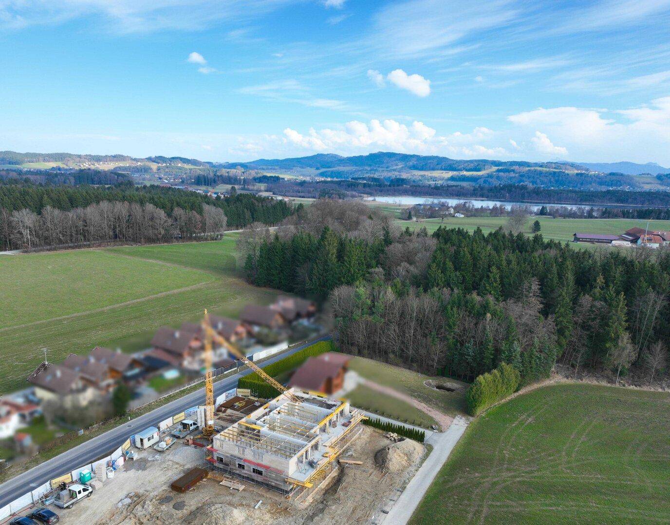 Luftaufnahme einer Baustelle mit Kran und Neubau in ländlicher Umgebung, umgeben von Feldern, Wäldern, einem See und Bergen unter blauem Himmel.