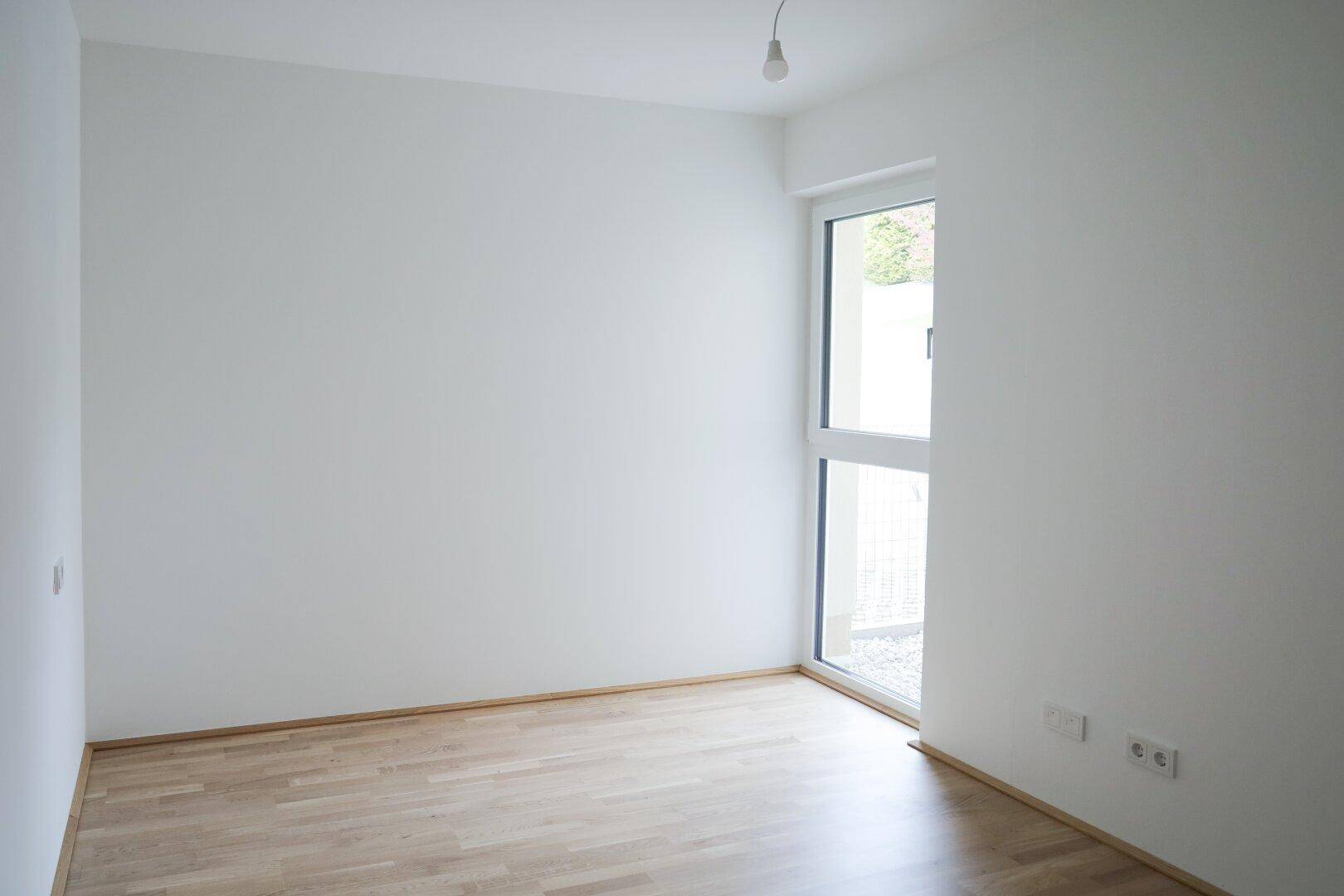 Heller, leerer Raum mit hellem Parkettboden und Zugang zu einem kleinen Außenbereich.