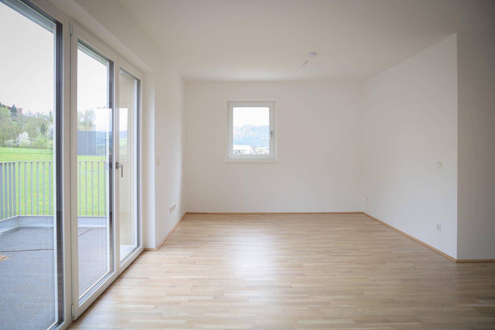 Helles Wohnzimmer mit Parkettboden und Zugang zu einem Balkon, umgeben von grüner Landschaft.