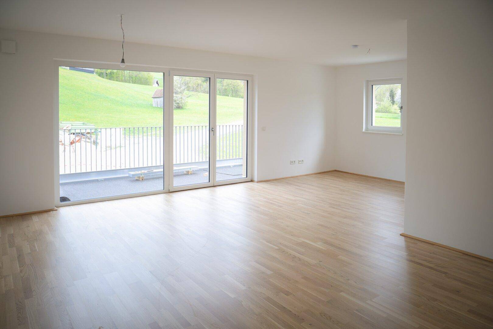 Geräumiges Wohnzimmer mit hellen Wänden, Holzboden und großem Fenster mit Blick ins Grüne.
