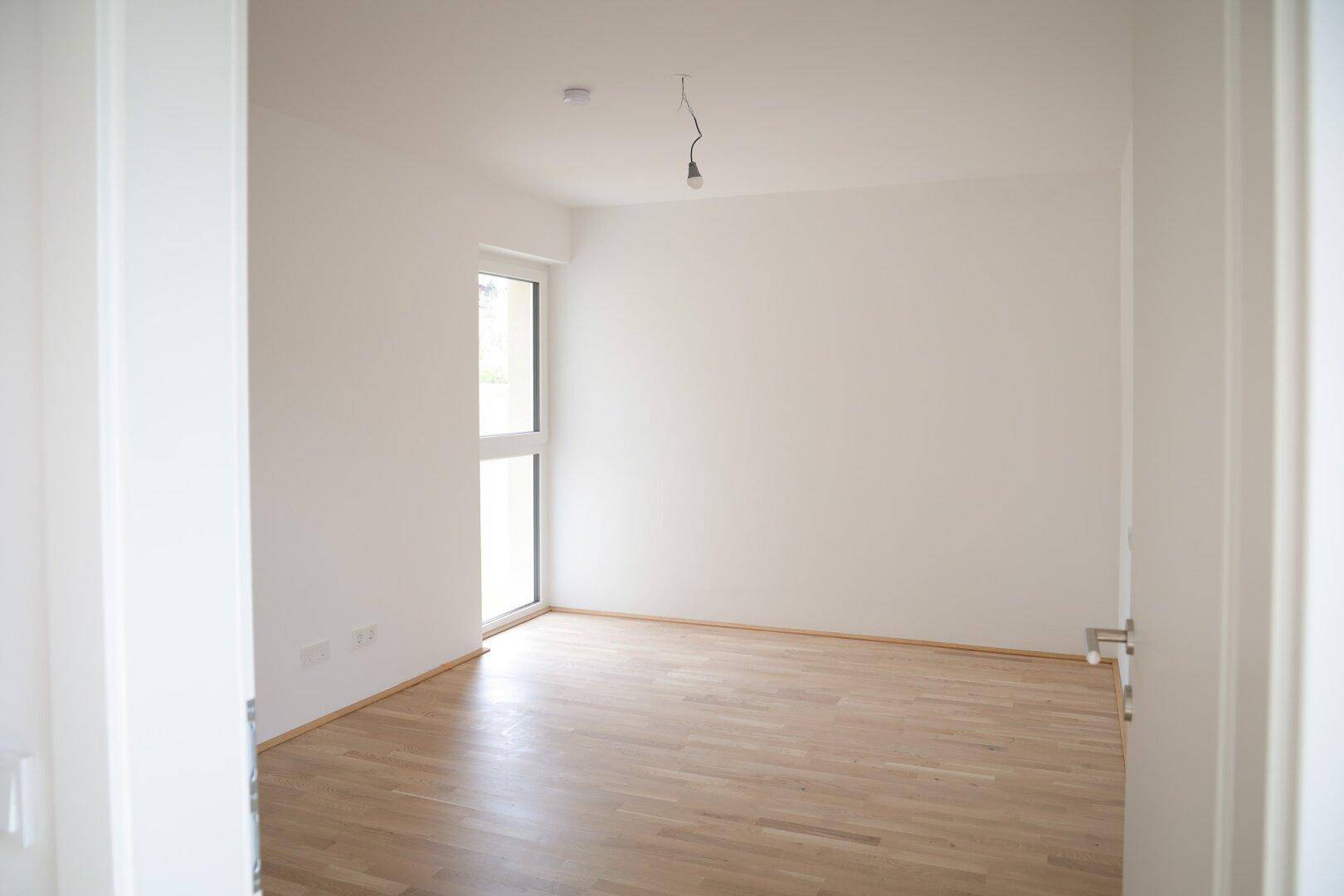 Leeres Schlafzimmer mit Parkettboden und einem großen Fenster, das viel Tageslicht hereinlässt.