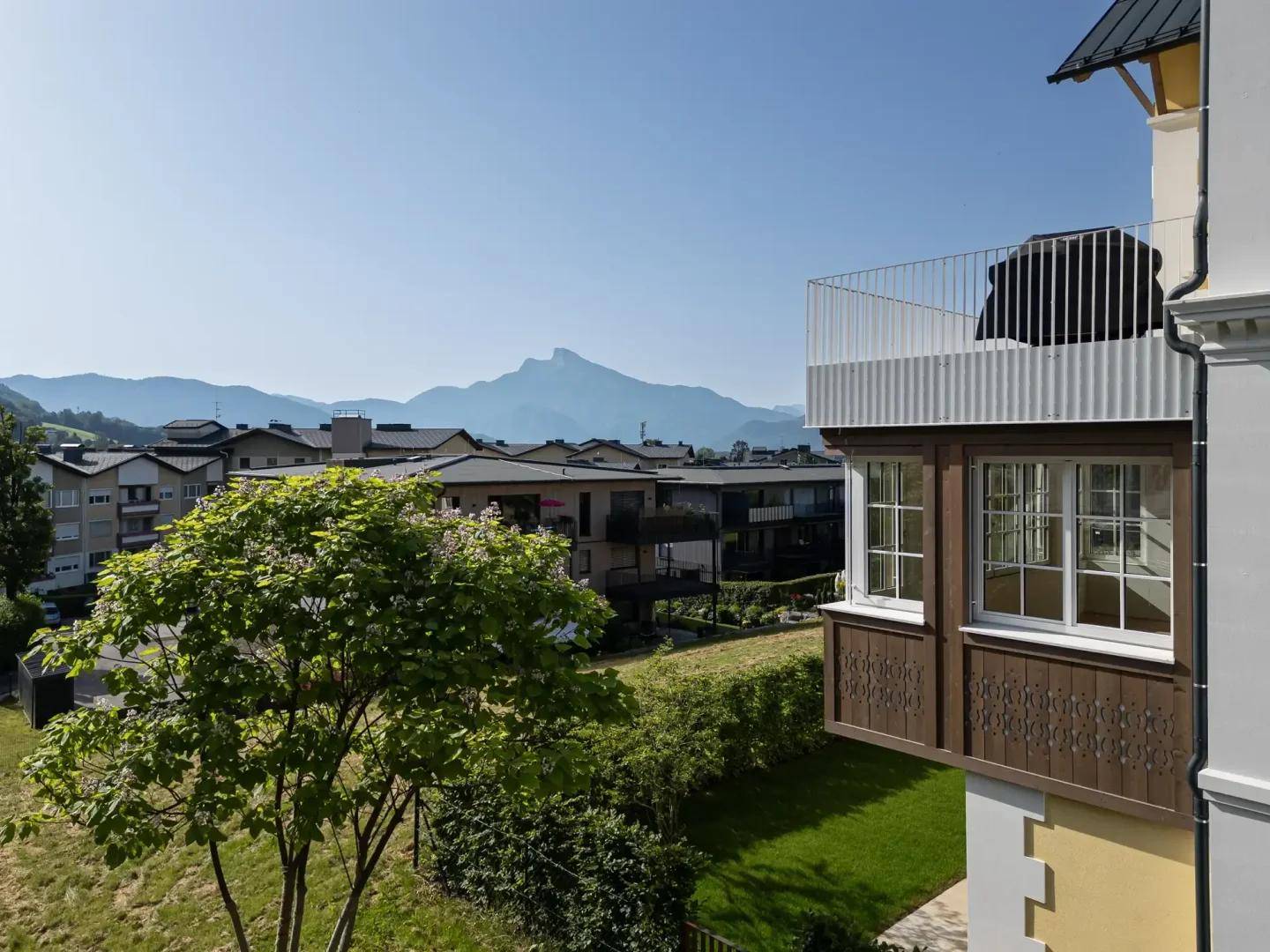 Balkon mit Bergblick und umliegender Wohnbebauung, umgeben von grüner Landschaft unter klarem Himmel.