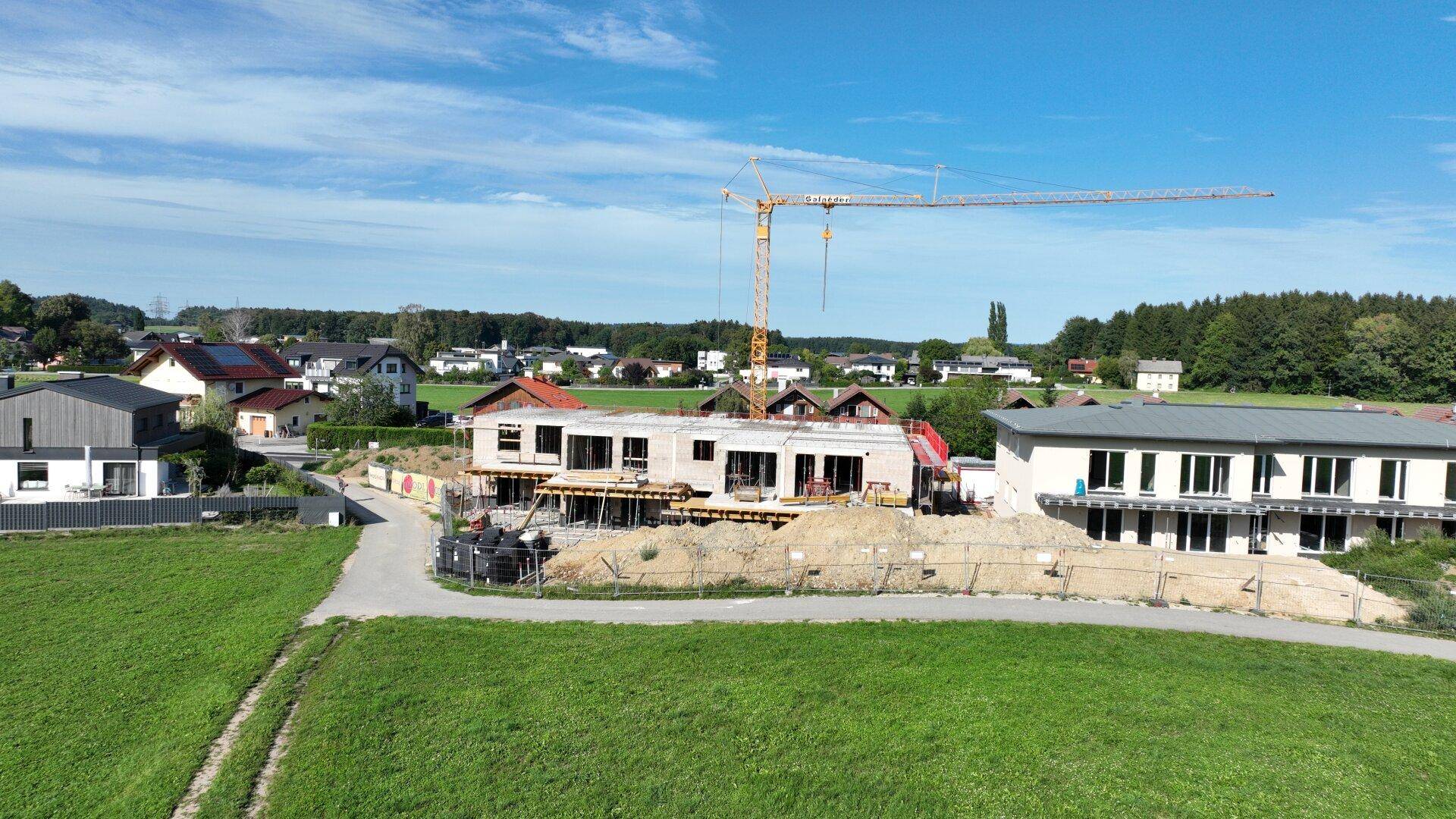 Baustelle eines neuen Gebäudes mit Kran, umgeben von Wohnhäusern und grünen Wiesen unter blauem Himmel.