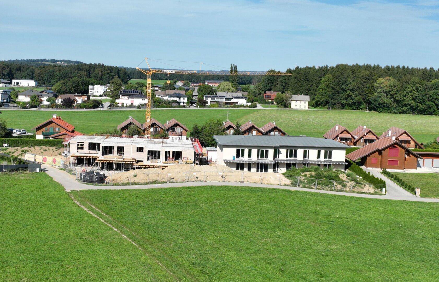 Luftaufnahme einer Baustelle mit zwei Gebäuden, umgeben von grünen Feldern und Wohnhäusern in ländlicher Umgebung.