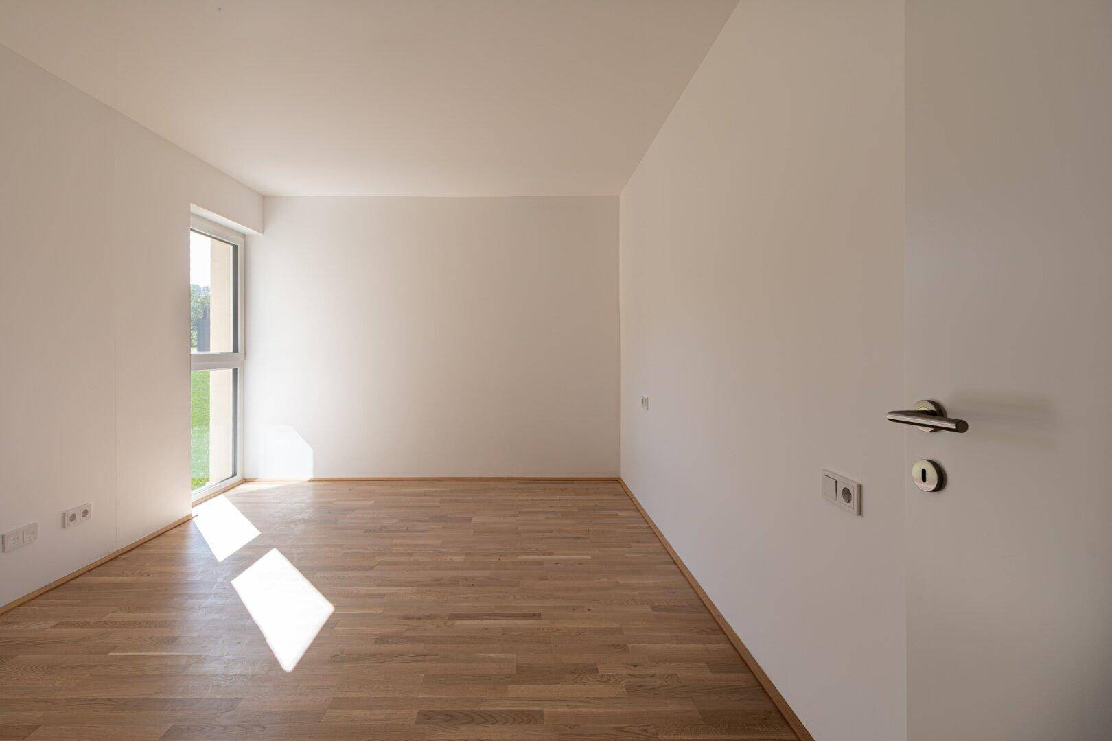 Helles Zimmer mit Parkettboden und einem Fenster, das viel Sonnenlicht hereinlässt.