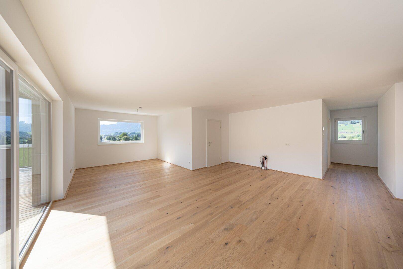 Geräumiger, heller Wohnbereich mit Holzboden und großen Fenstern, die viel Tageslicht hereinlassen.