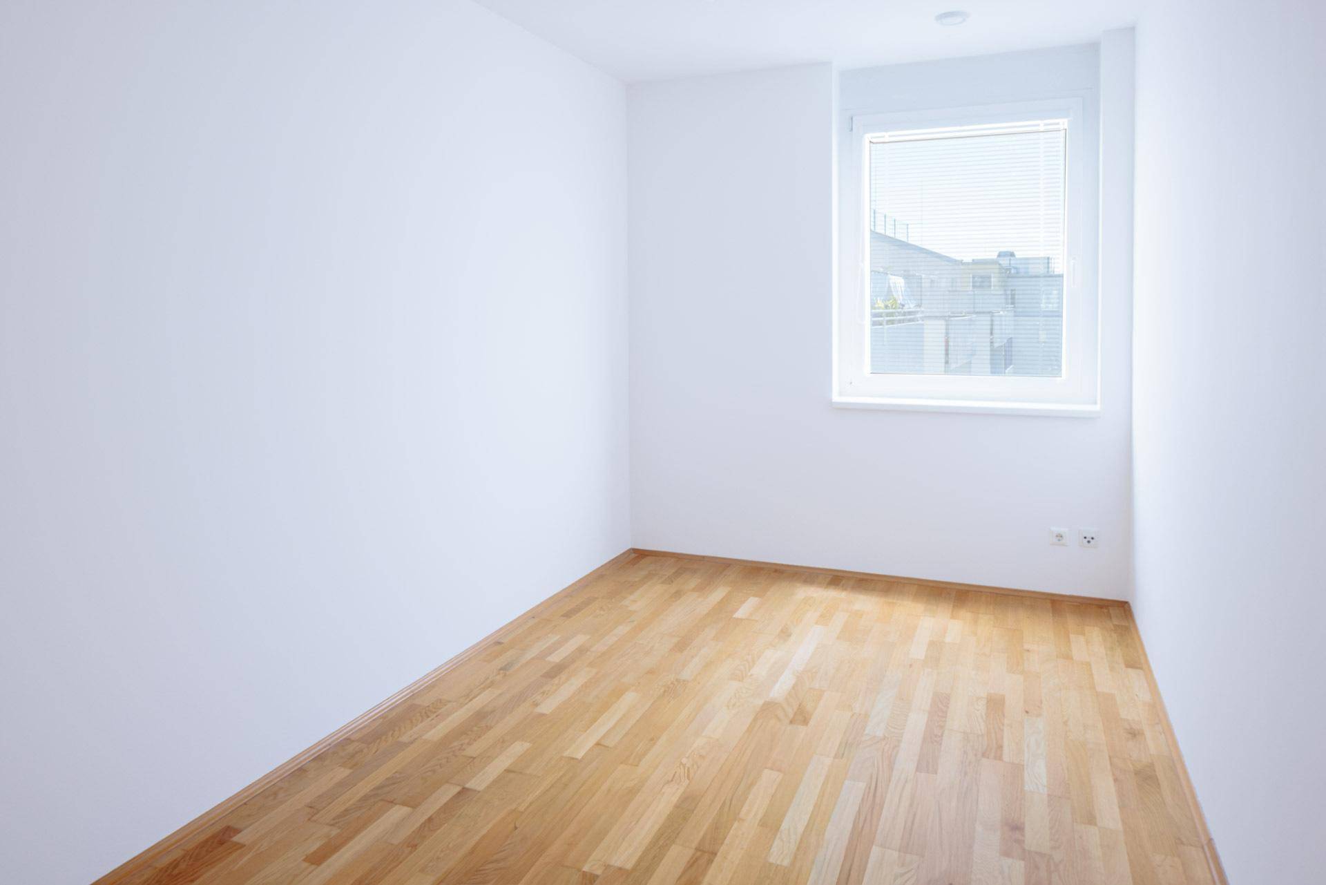 Ruhiges Schlafzimmer mit hellem Parkettboden und einem Fenster mit Jalousien, ideal zum Entspannen.
