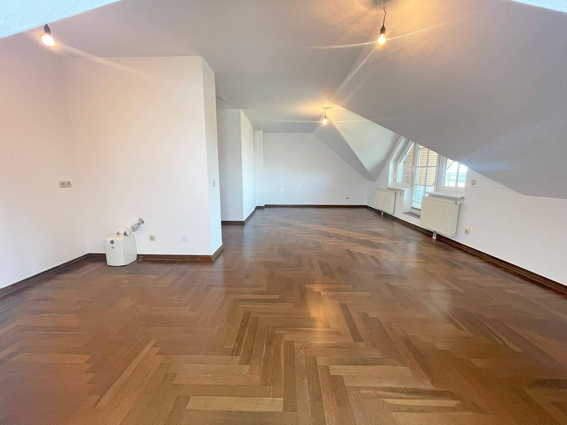 Weitläufiger Wohnbereich im Dachgeschoss mit klassischem Fischgrätparkett und weiß gestrichenen Wänden.