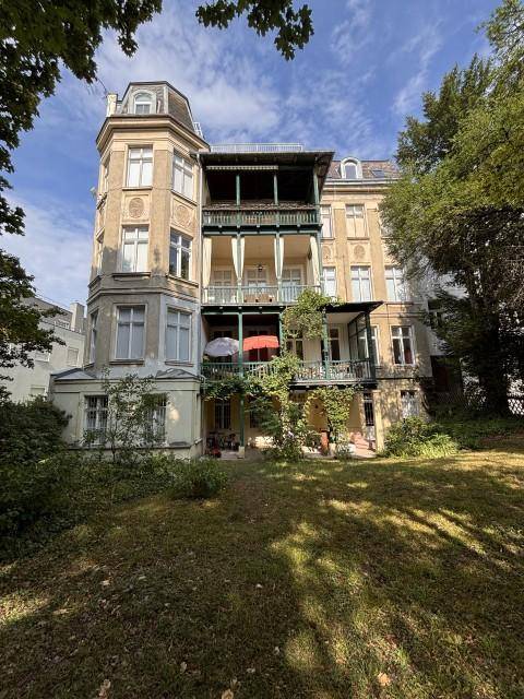 Historisches Gebäude mit mehreren Balkonen und einem großen, grünen Gartenbereich im Vordergrund.