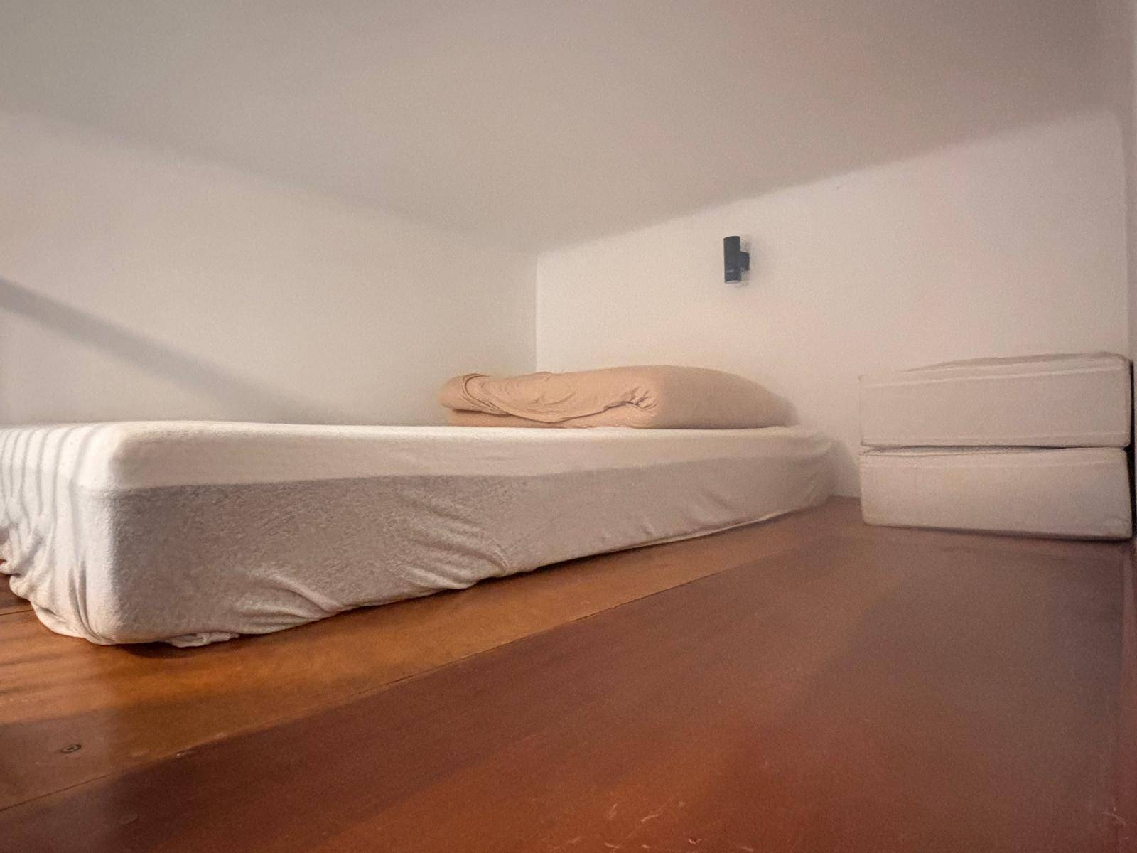 Schlafbereich auf einer erhöhten Holzplattform mit einfacher Matratze und niedriger Deckenhöhe.