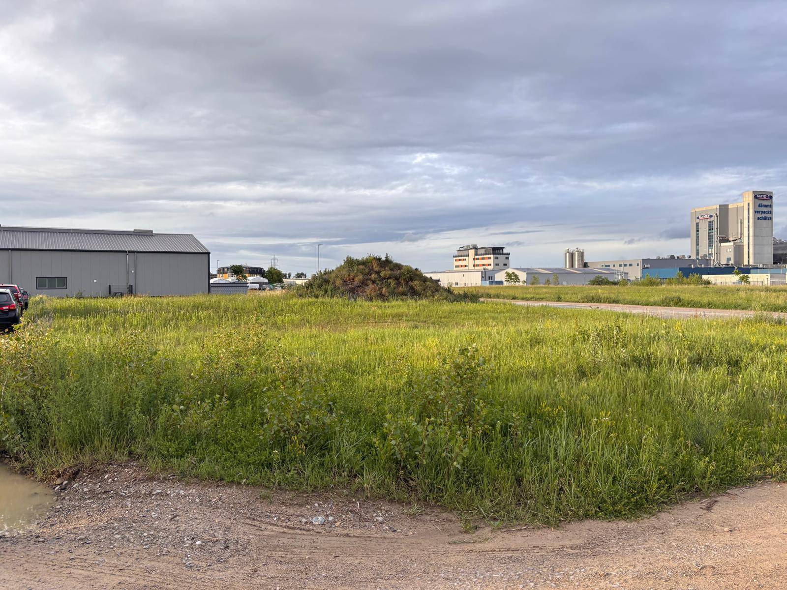 Weite Grasfläche mit Erdhügel und mehreren Industriegebäuden im Hintergrund unter einem bewölkten Himmel.