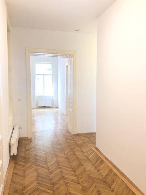 Langer Flur mit Fischgrätparkett, der zu einem hellen Raum mit Fenster und Heizkörper führt.