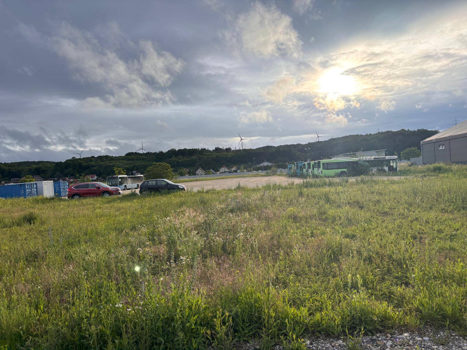 Grünfläche mit parkenden Fahrzeugen und Bussen, die Sonne scheint durch die Wolken, im Hintergrund Windräder.
