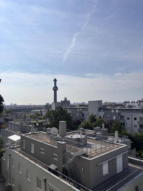 Weitläufiger Stadtblick über Dächer und Gebäude unter blauem Himmel, mit einem markanten Turm in der Ferne.