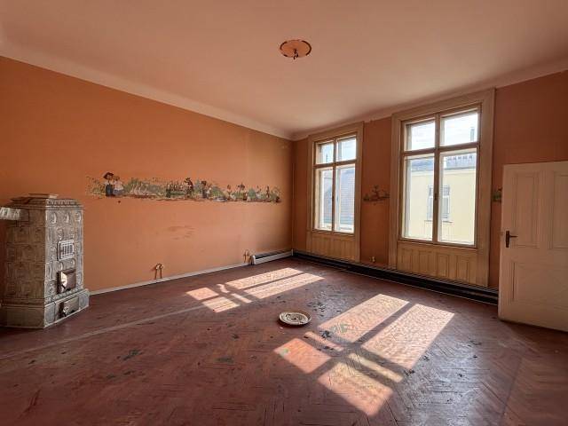 Geräumiges Zimmer mit Kachelofen, Fischgrätparkett und zwei großen Fenstern, die viel Licht hereinlassen.