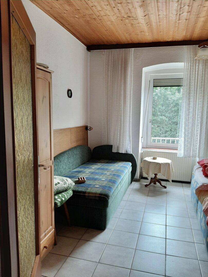 Schlafzimmer mit Einzelbett, Fliesenboden, Holzdecke und Fenster mit Blick ins Grüne.