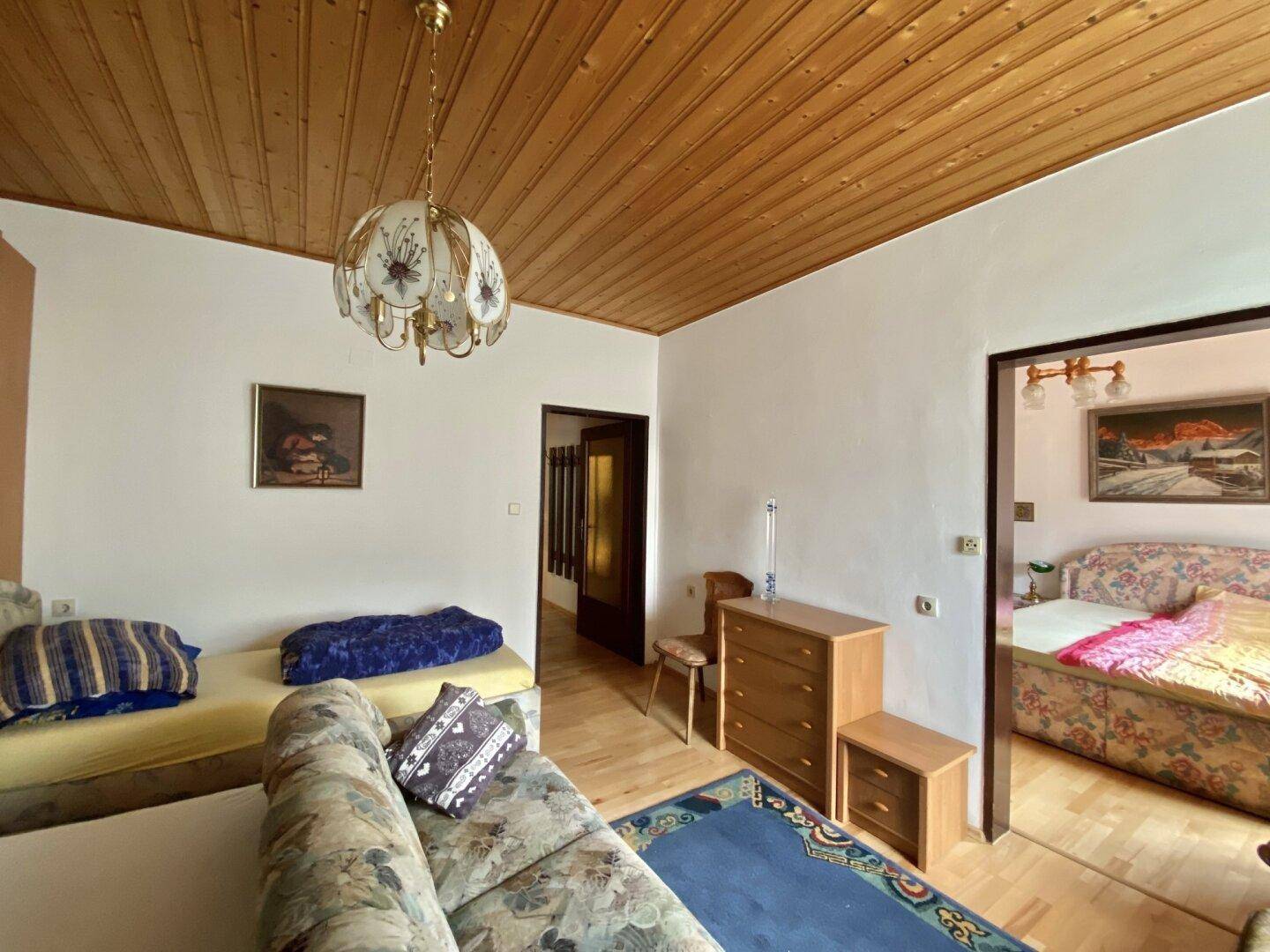 Wohnzimmer mit Holzdecke, Teppichboden und traditionellen Möbeln, angrenzend an ein Schlafzimmer.