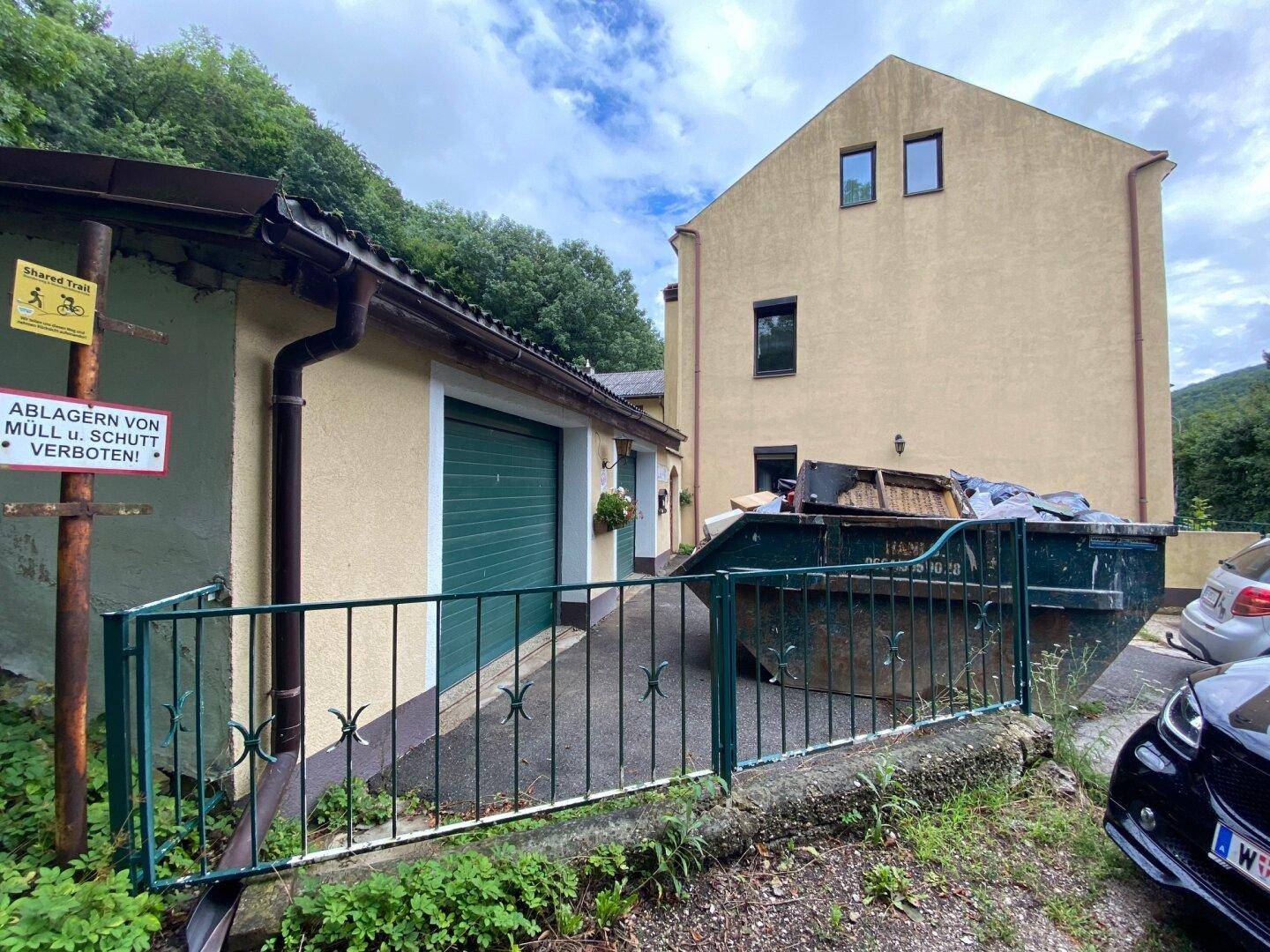 Seitenansicht des Gebäudes mit Garage, Müllcontainer und bewachsenem Hang.