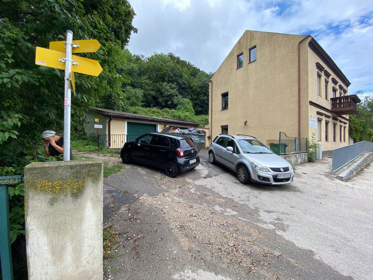Ansicht des Gebäudes mit Garagen, geparkten Autos und Wegweisern im Vordergrund.