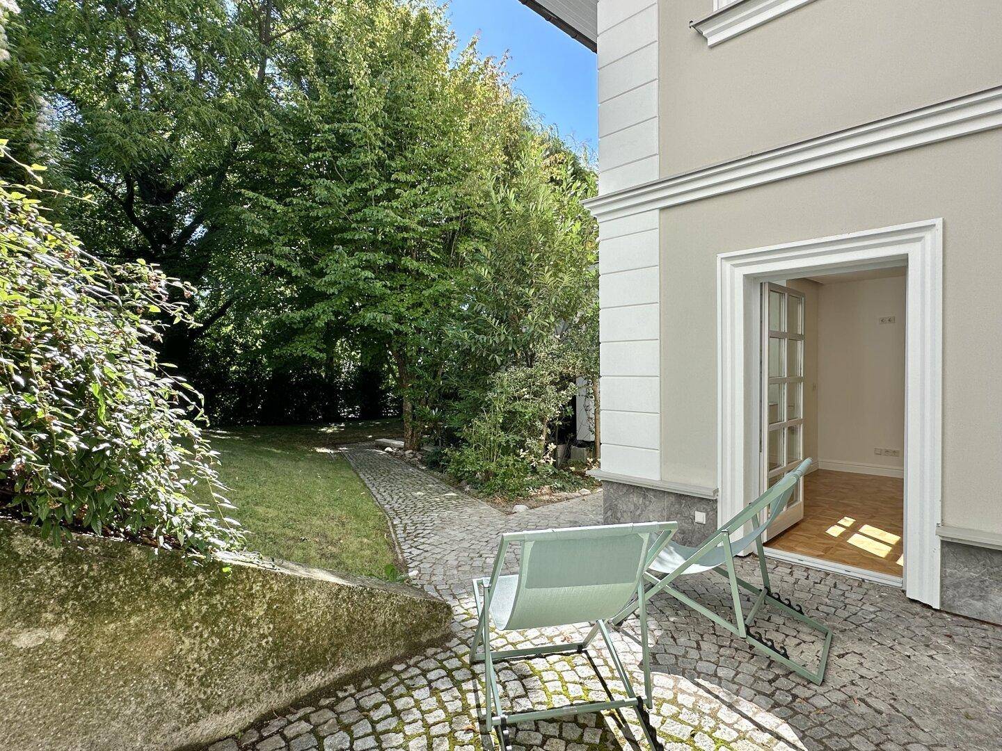 Sonniger Gartenbereich mit gepflastertem Weg, Rasenfläche und zwei Liegestühlen vor dem Haus.