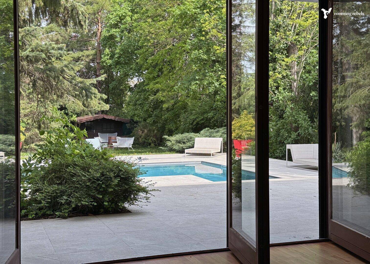 Blick aus dem Innenraum durch große Glasfronten auf den Pool und den grünen Gartenbereich.