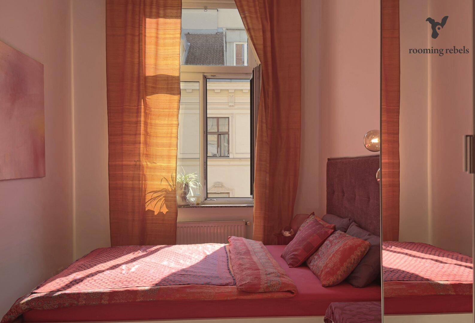 Gemütliches Schlafzimmer mit pinkfarbenem Bett, orangefarbenen Vorhängen und Blick auf die Stadt durch das Fenster.