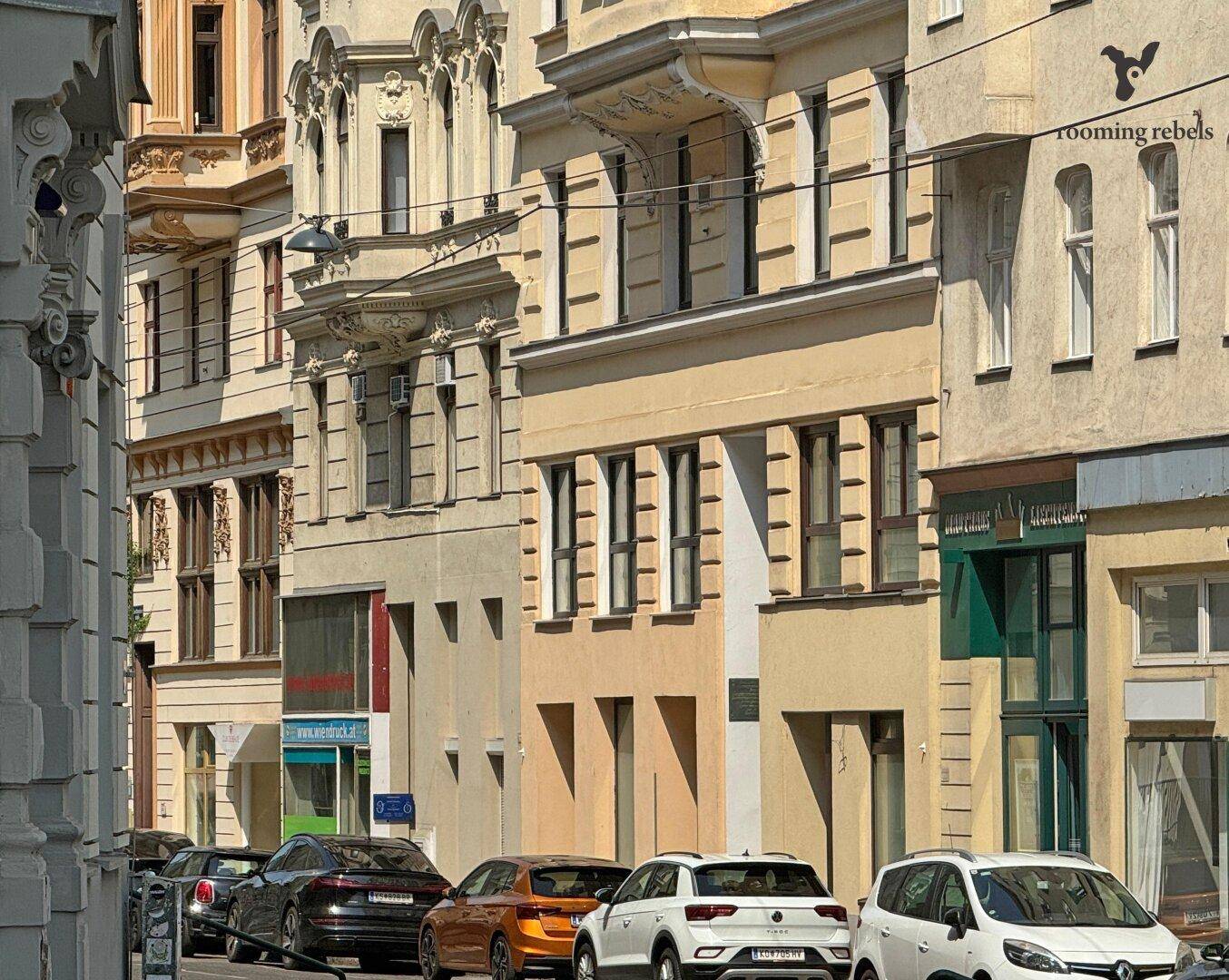 Historische Gebäudezeile mit reich verzierten Fassaden und parkenden Autos auf der Straße.