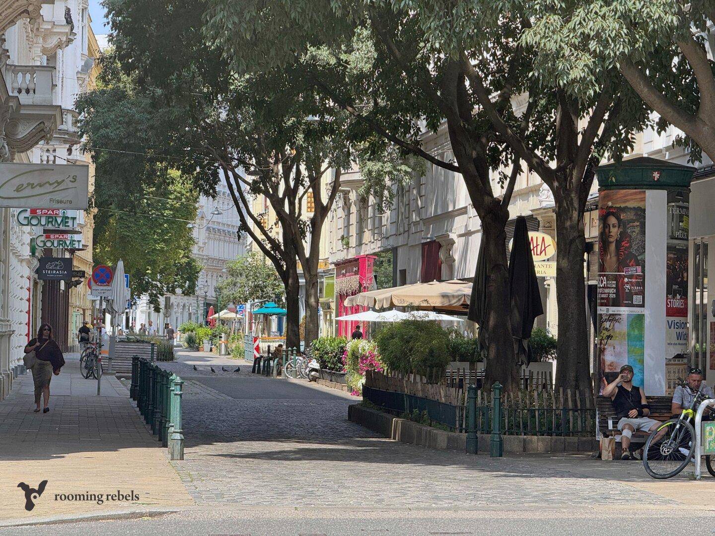 Ruhige Stadtstraße mit Bäumen, Geschäften und Außensitzbereichen unter blauem Himmel.