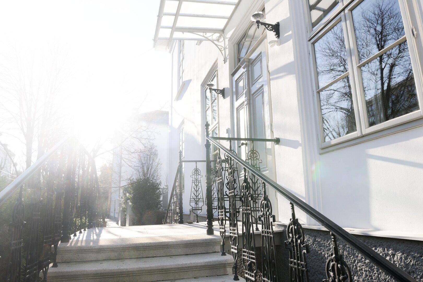Sonniger Eingangsbereich eines eleganten Gebäudes mit Treppe und schmiedeeisernem Geländer.