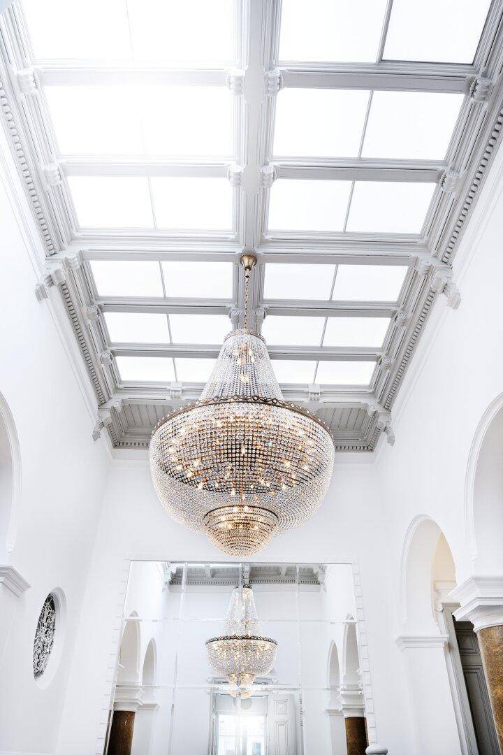 Imposanter Kronleuchter hängt von einer hohen Decke mit Glasdach in einem eleganten Treppenhaus.