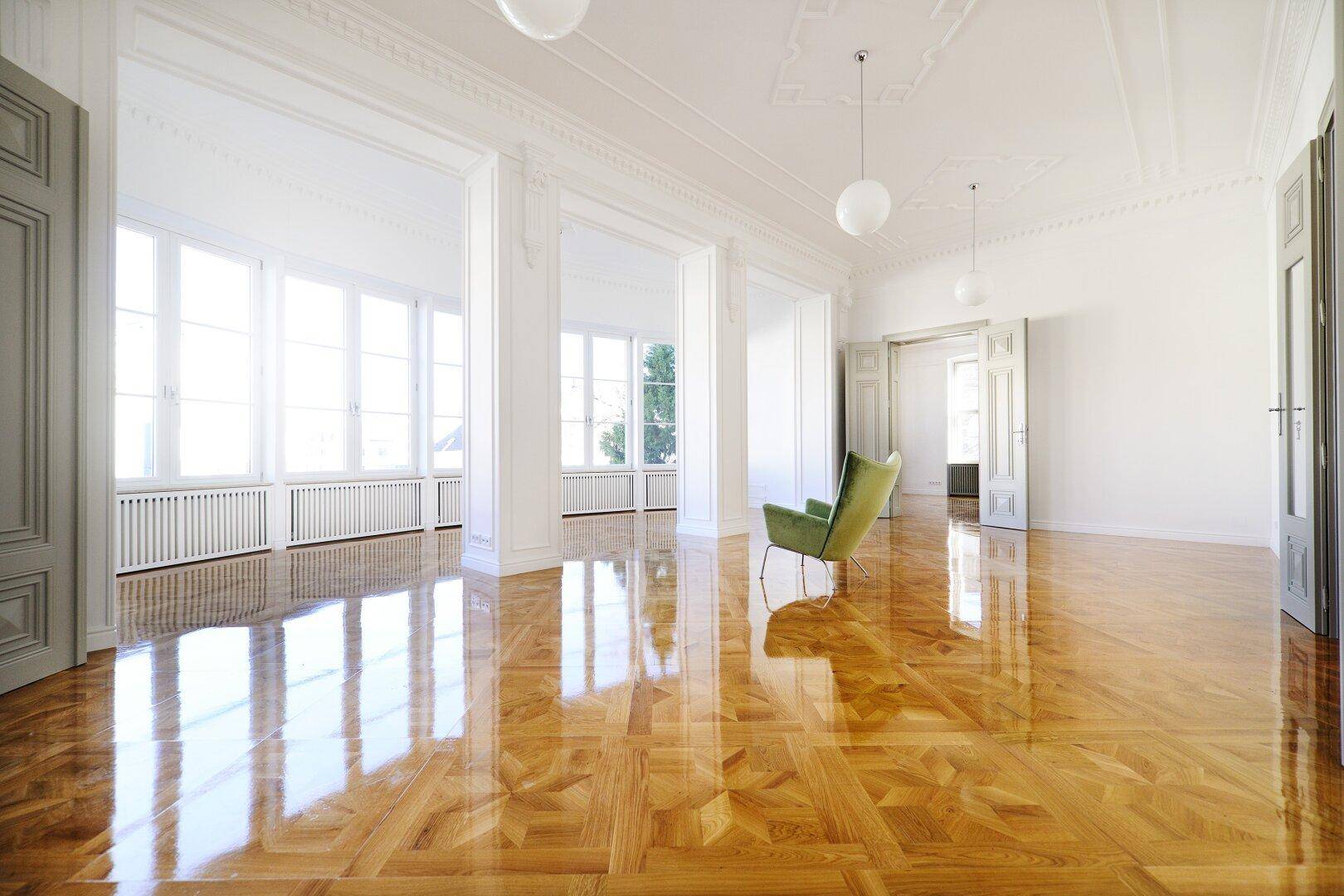 Großer, heller Wohnraum mit glänzendem Parkettboden, hohen Decken und großen Fenstern.