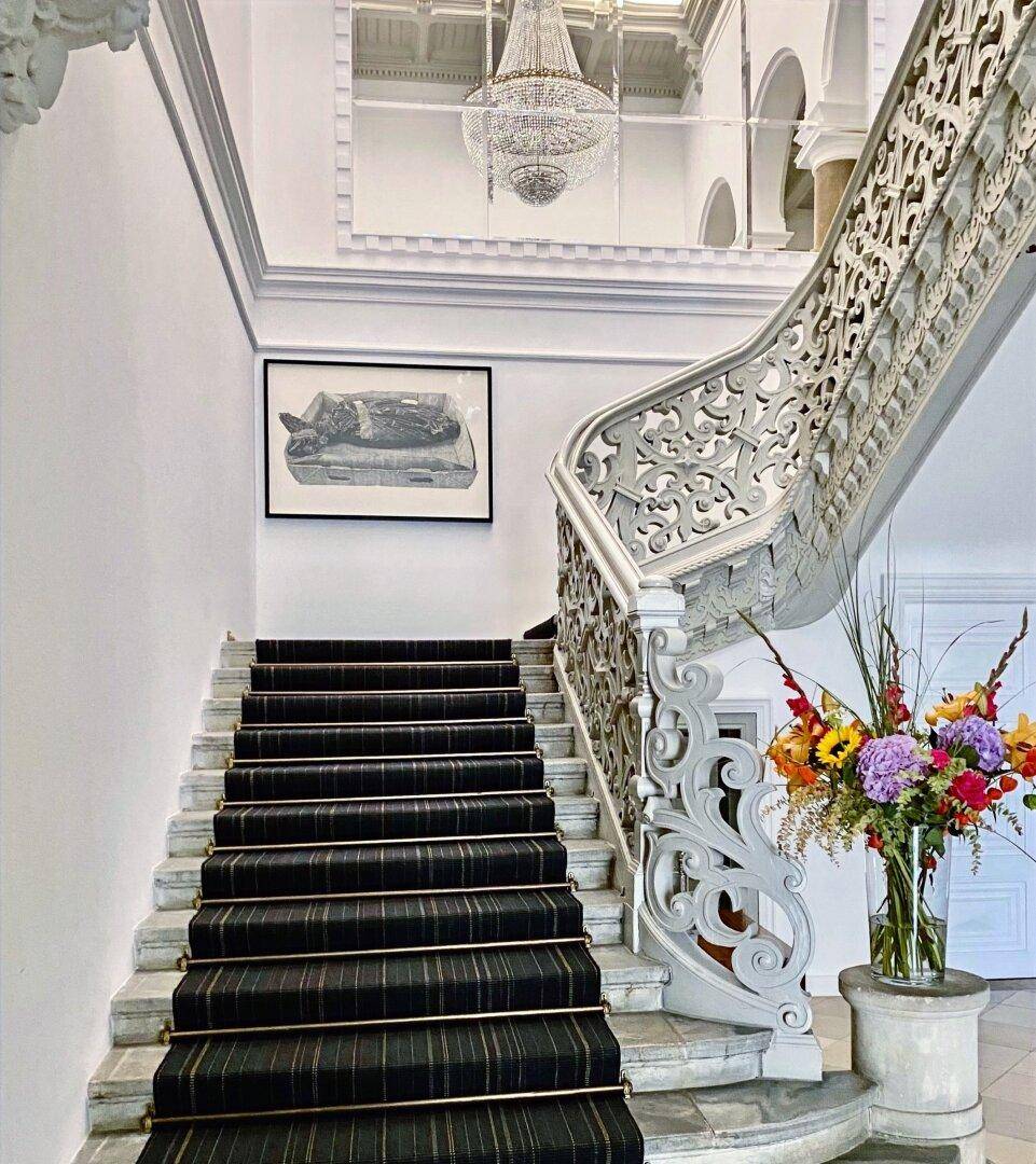 Elegantes Treppenhaus mit schwarzem Teppich, kunstvollem Geländer und einem Blumenarrangement auf einem Podest.