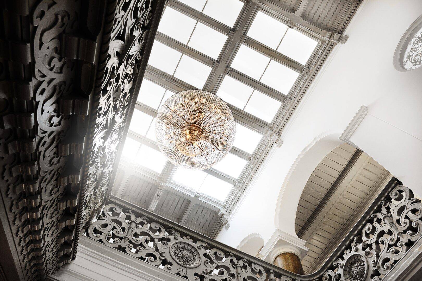Elegantes Treppenhaus mit kunstvollem Geländer, großem Kronleuchter und natürlichem Licht durch das Oberlicht.