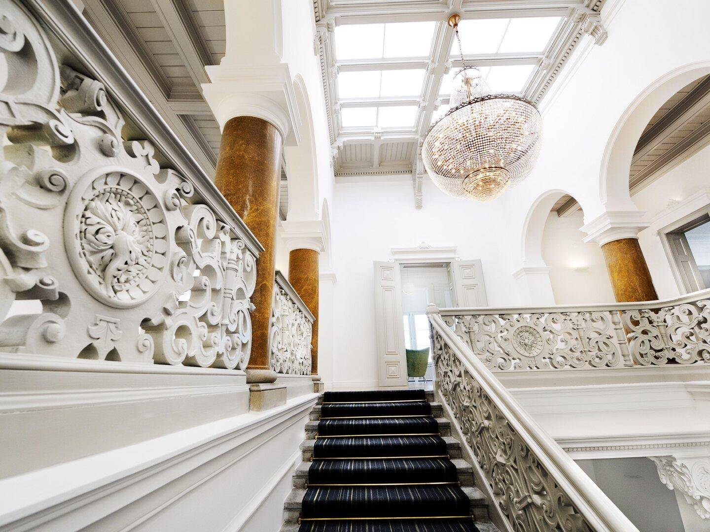 Majestätisches Treppenhaus mit schwarzem Teppich auf den Stufen, kunstvollem Geländer und einem Kronleuchter.