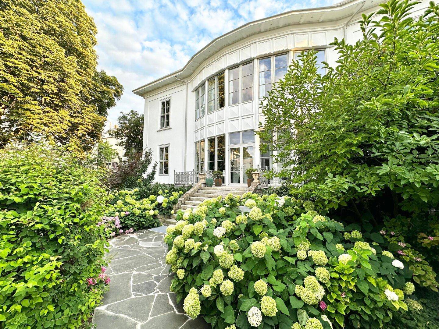 Elegante weiße Villa mit großen Fenstern und einem üppigen, gepflegten Garten unter blauem Himmel.