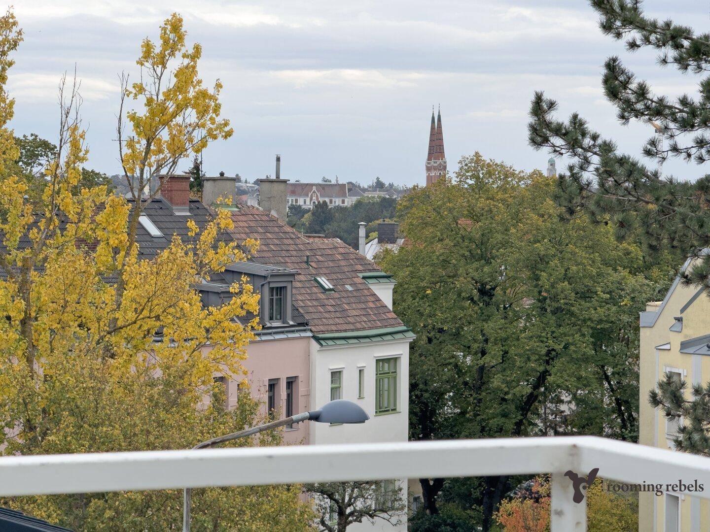Ein Blick über Dächer und Bäume auf eine Stadtlandschaft mit einem Kirchturm in der Ferne.